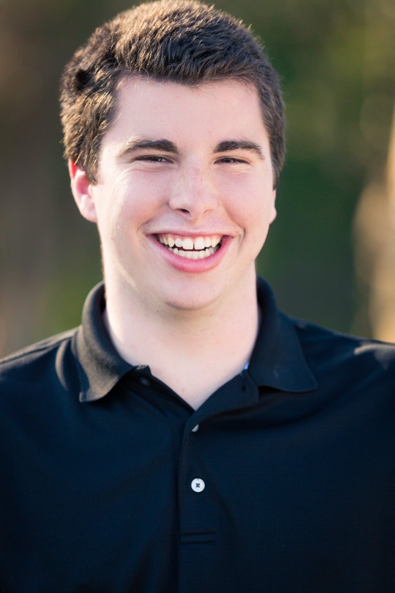 High school senior in a navy polo laughing outdoors against soft green foliage