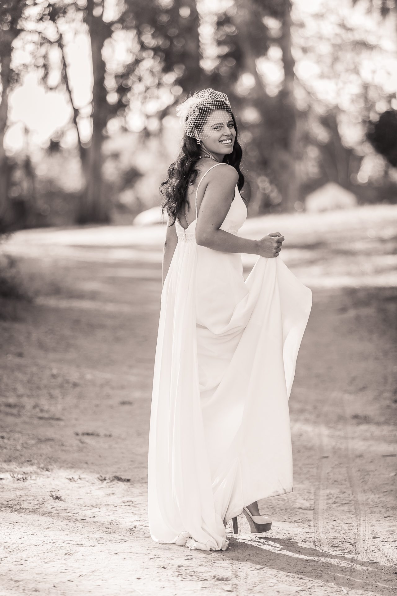 Sepia portrait of a bride in a birdcage veil walking a dirt path, holding her dress and smiling back over her shoulder