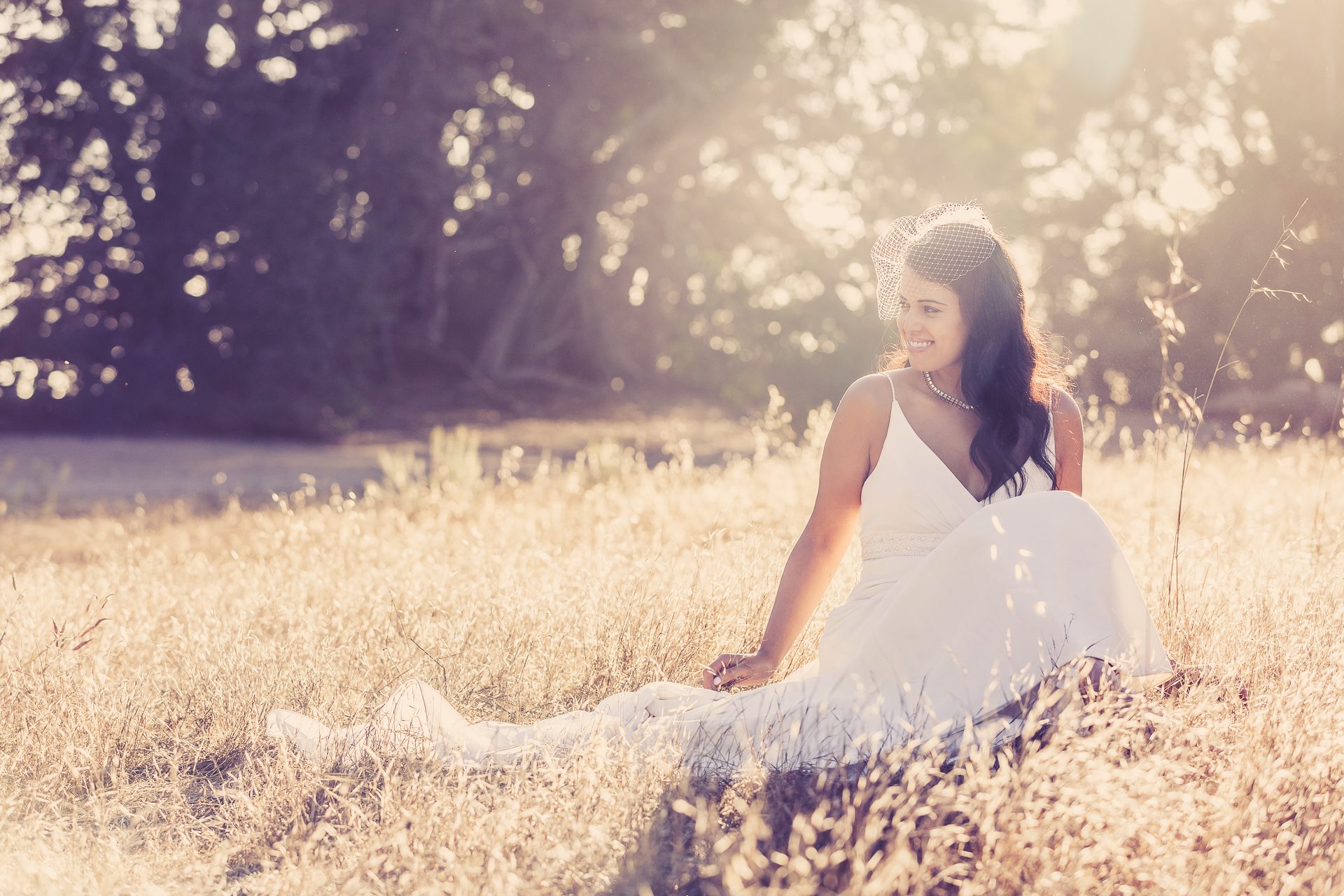 Bride in a birdcage veil sitting in golden dry grass as the sun flares through the trees