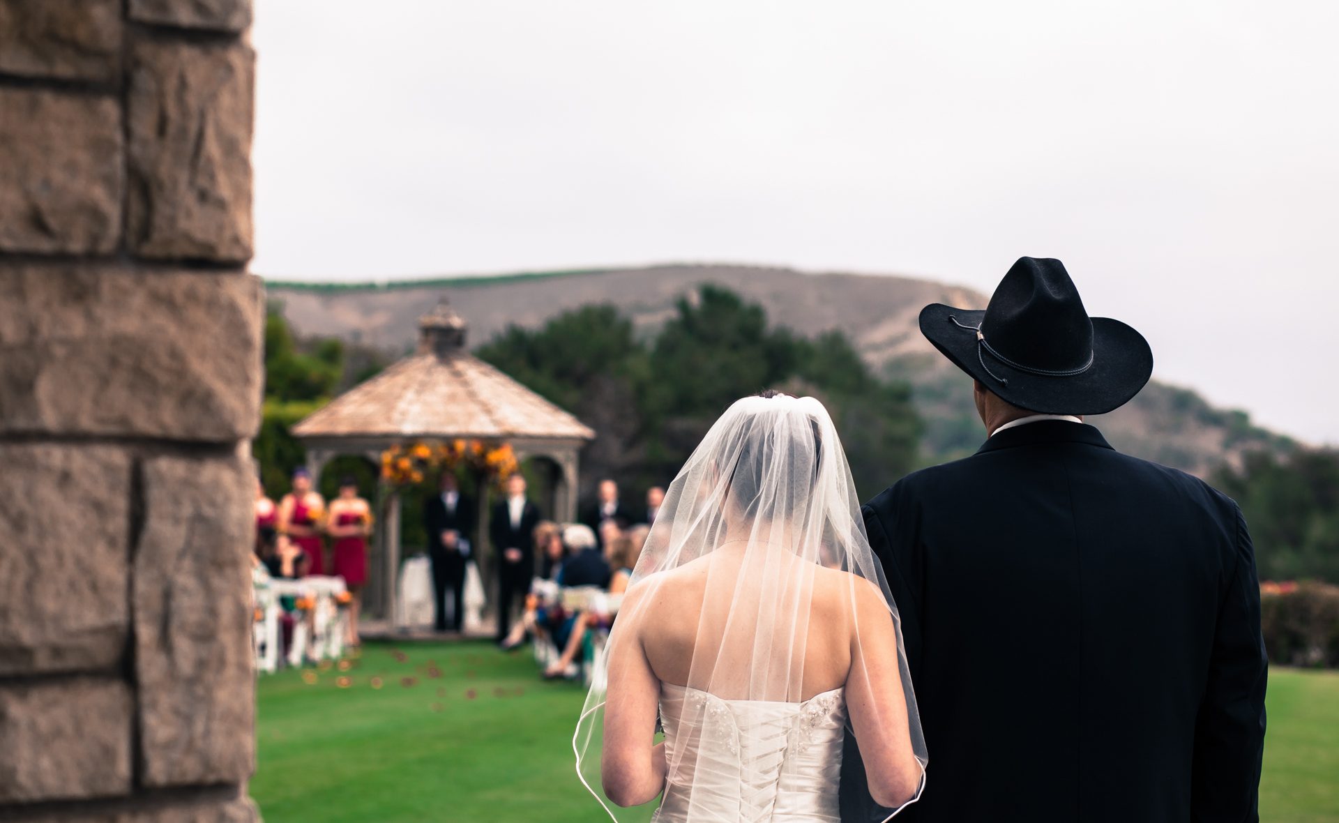 Bride in her veil walking arm in arm with a man in a black cowboy hat toward a garden gazebo ceremony