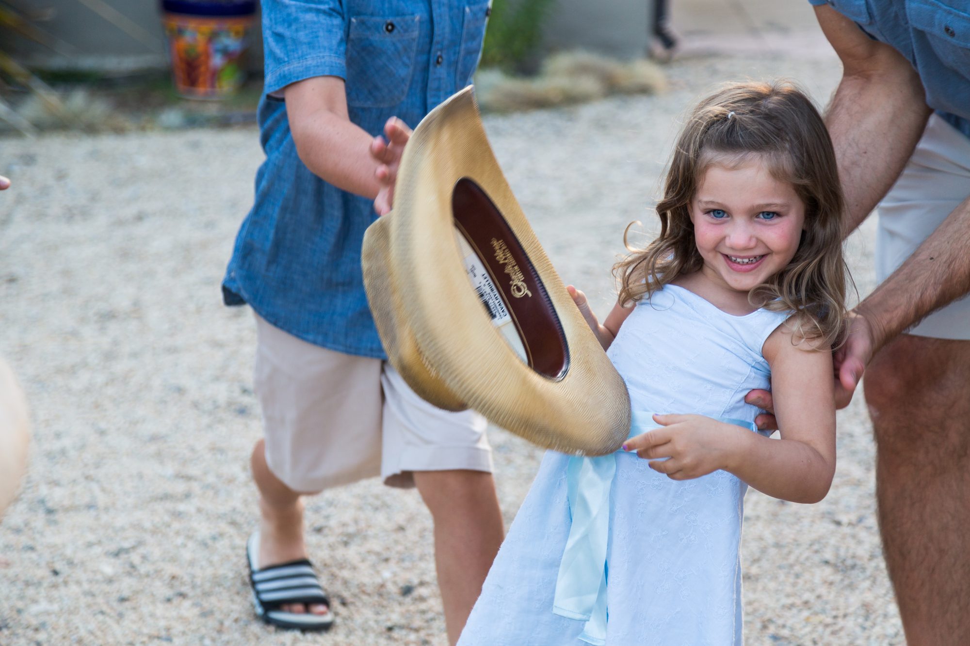 Little girl in a light-blue dress laughing as a straw cowboy hat is tossed past her at a backyard party
