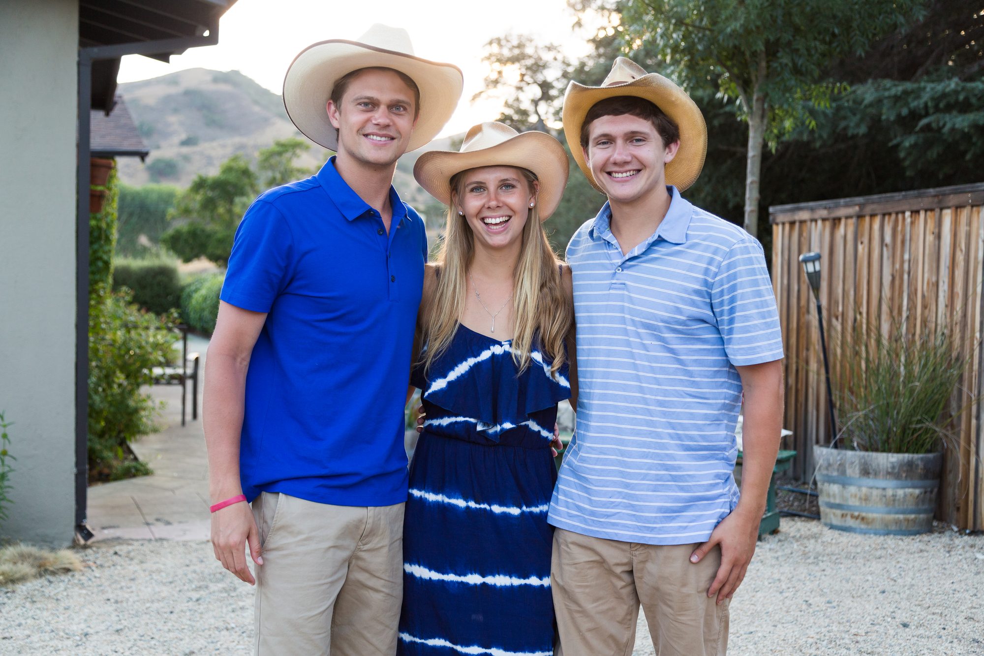 Three young adults in straw cowboy hats smiling together at a backyard party