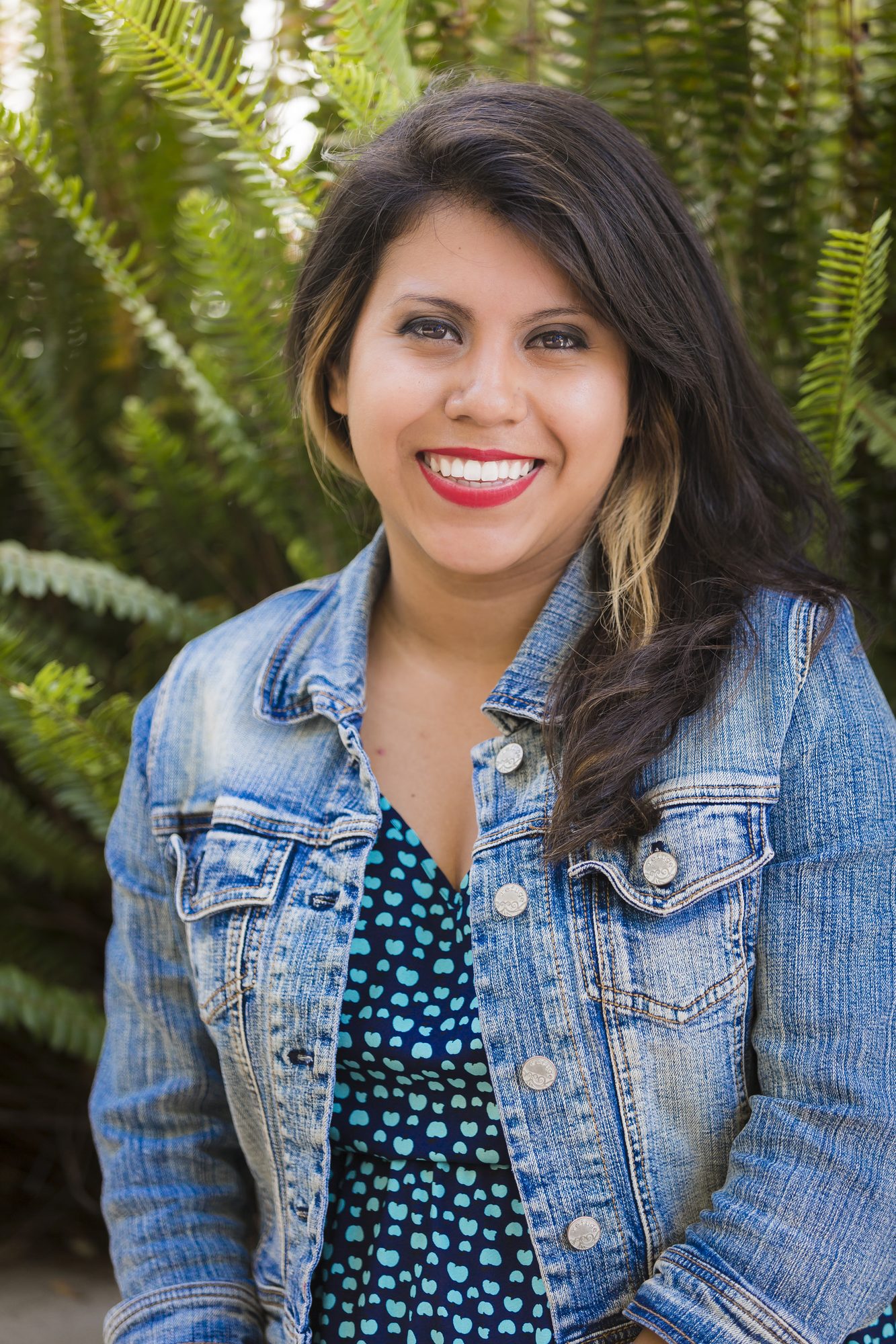 Smiling woman in a denim jacket and red lipstick with ferns behind her