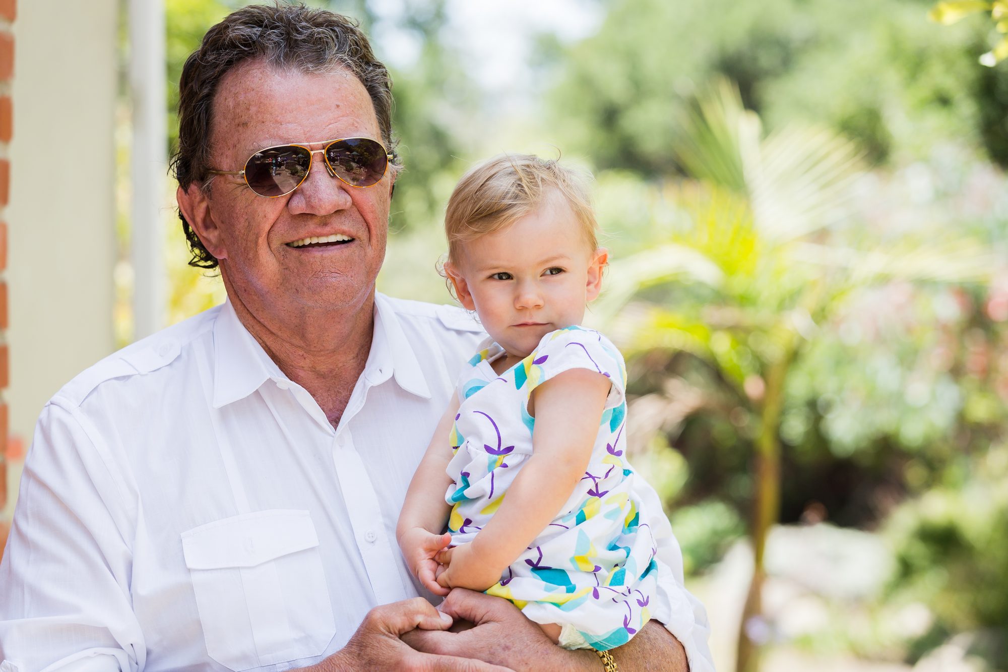 Grandfather in aviator sunglasses holding his blonde granddaughter in a floral dress