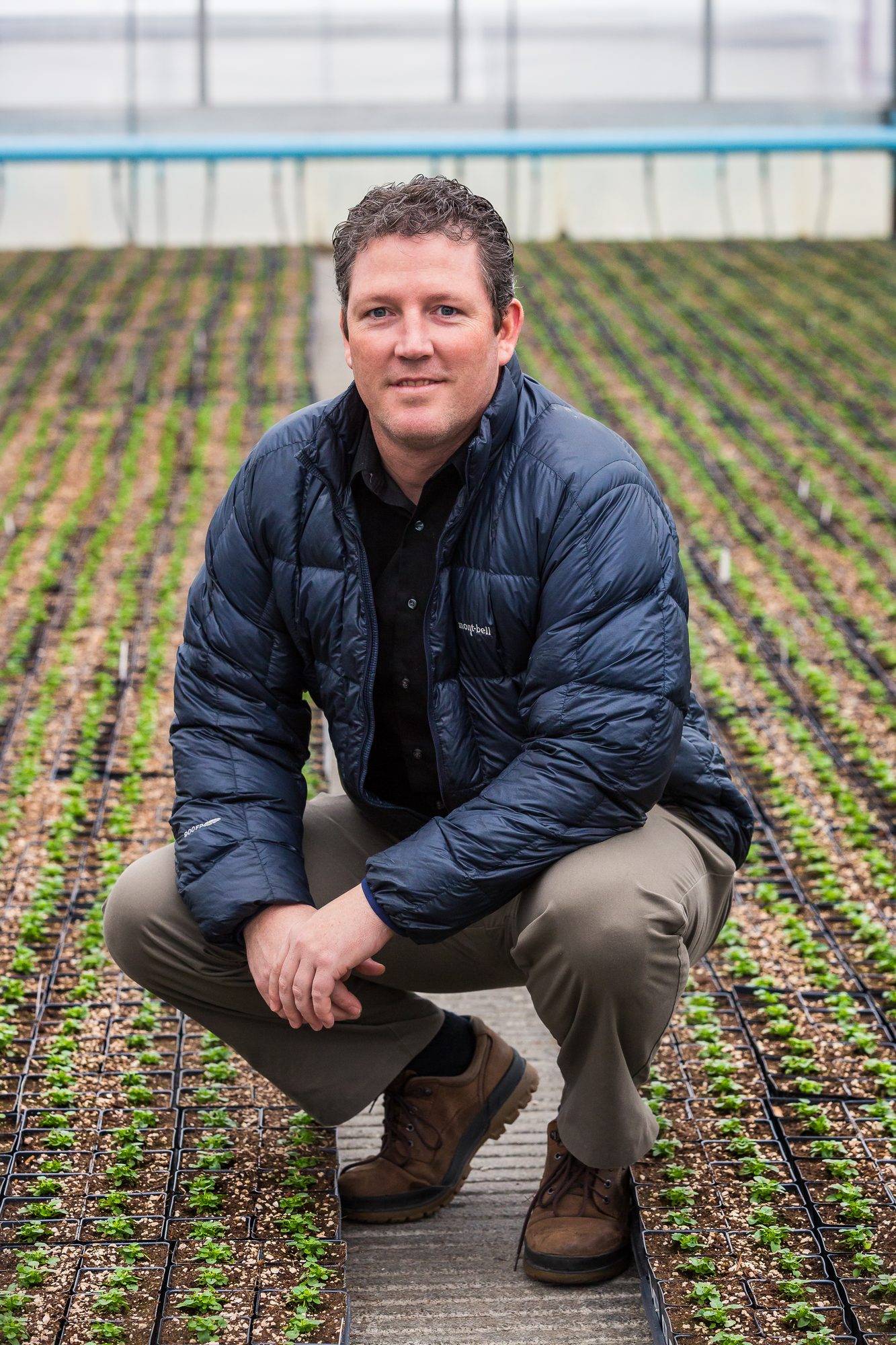 Grower in a quilted jacket crouching between rows of seedlings in a greenhouse