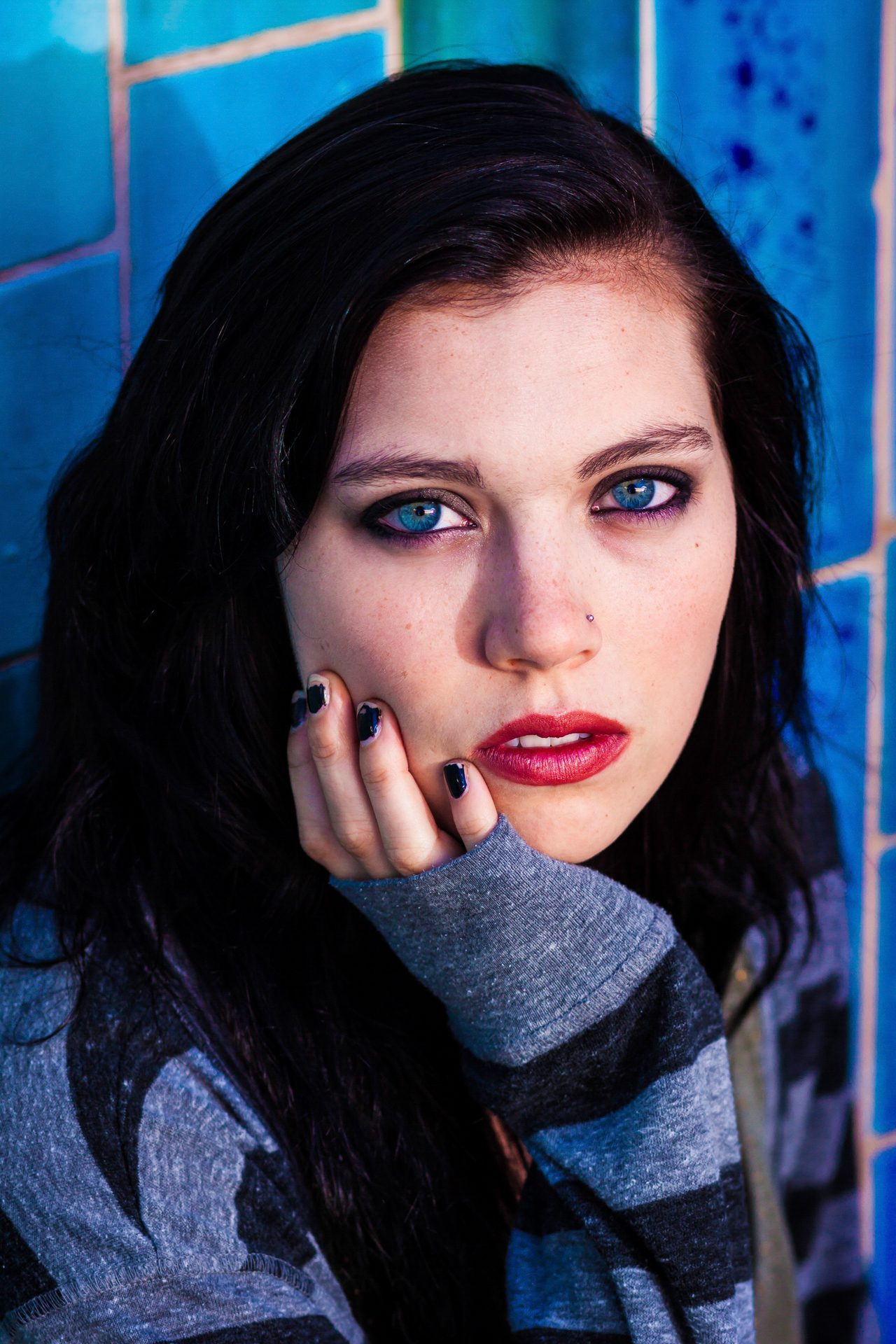 Dark-haired young woman with blue eyes and red lips, hand resting on her cheek, against blue tile