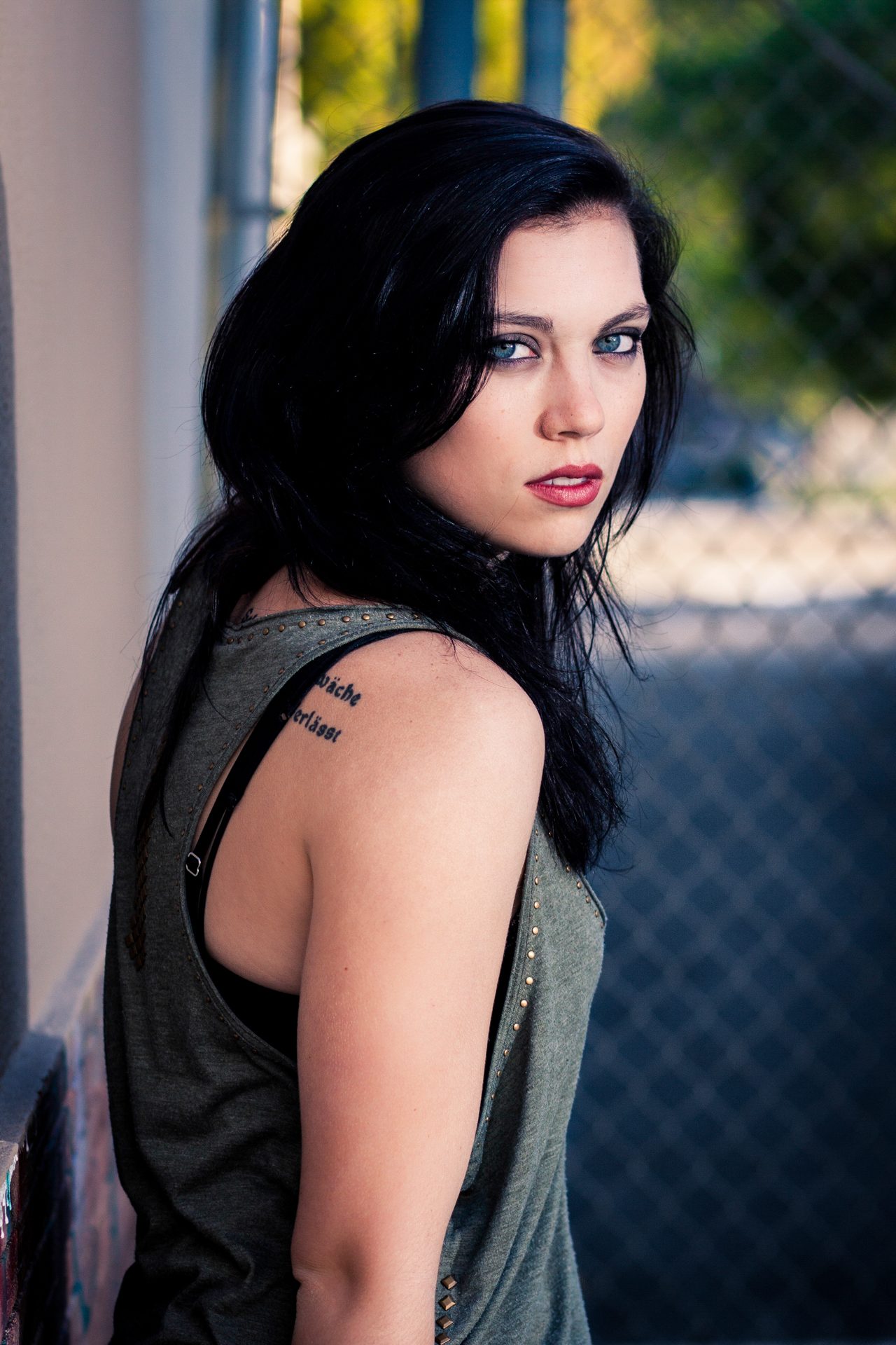Dark-haired woman with blue eyes looking back over a tattooed shoulder, warm foliage behind