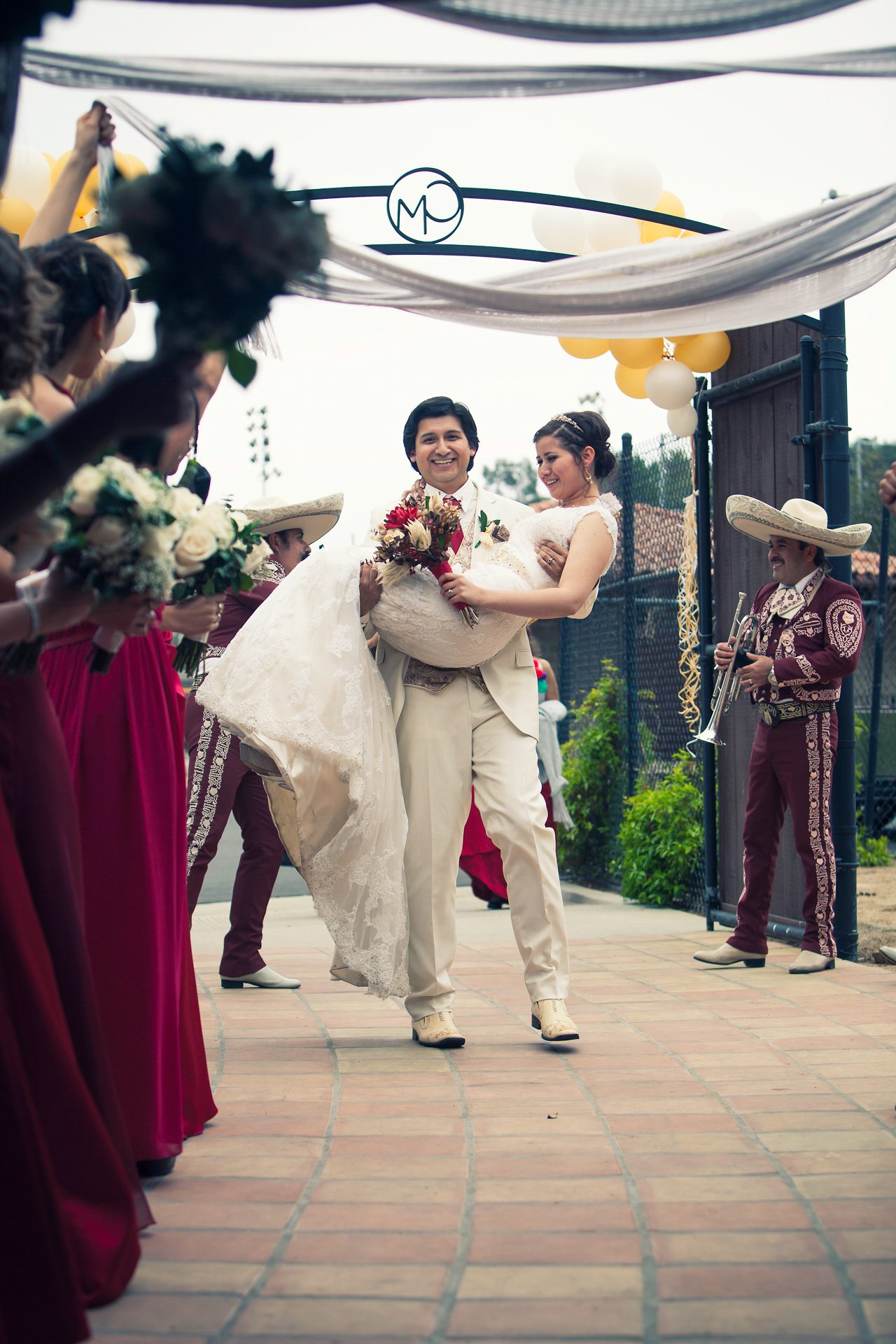 Groom carrying his bride beneath a flower arch as mariachi musicians play