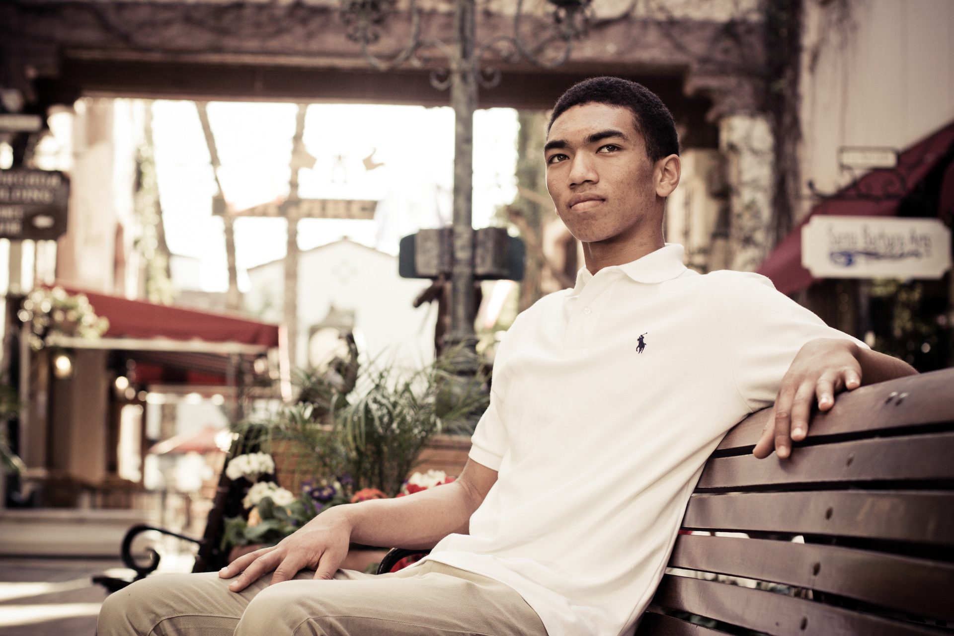 Senior in a white polo seated on a wooden bench in a sunlit plaza