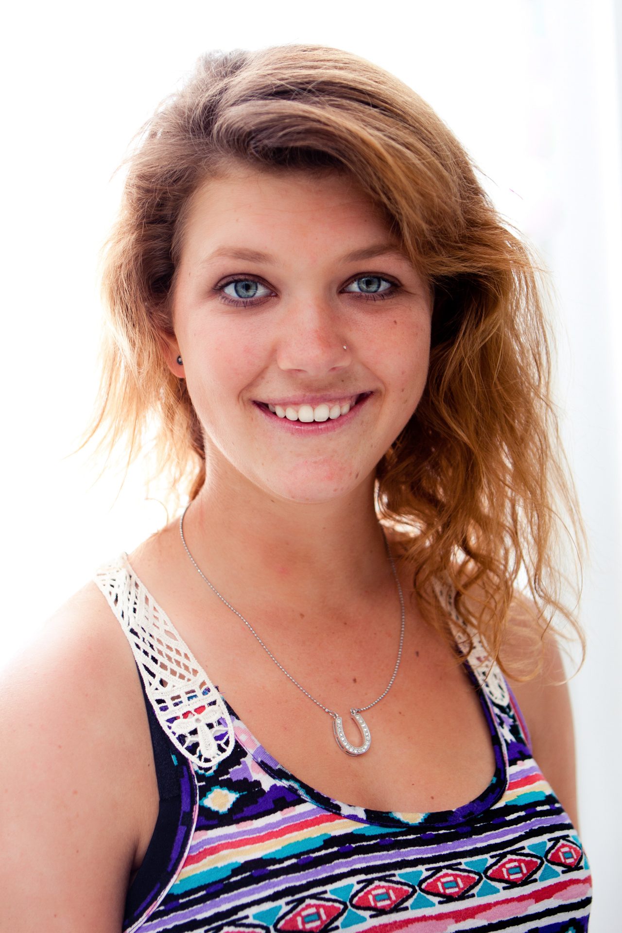 Smiling young woman with wavy auburn hair and blue eyes in a patterned tank top, bright daylight behind