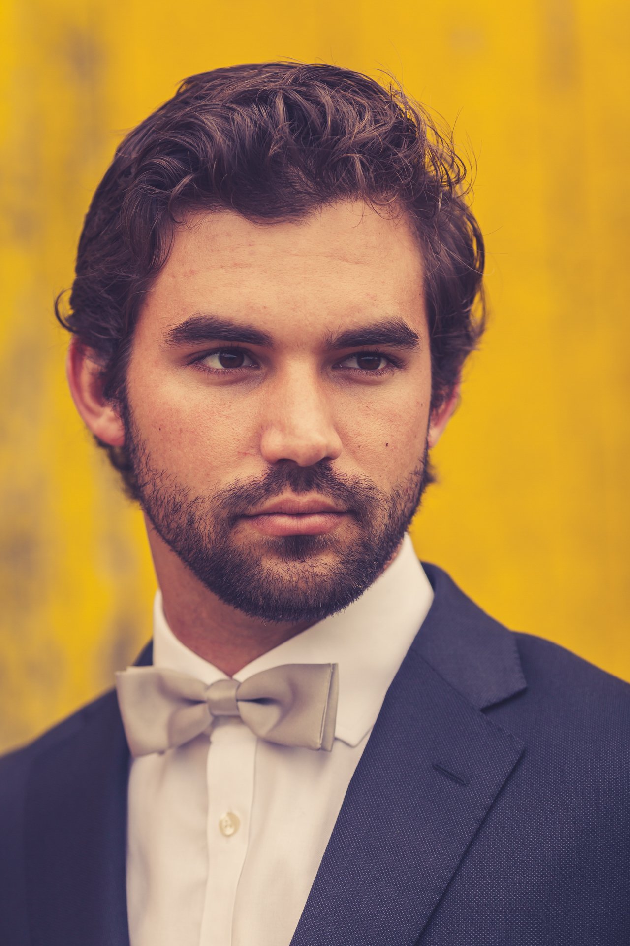Editorial portrait of a bearded man in a navy suit and pale bow tie against a weathered yellow wall