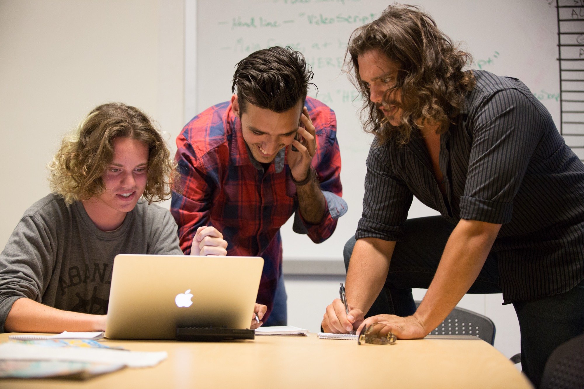 Three student journalists gathered around a laptop, one laughing on the phone, in a newsroom