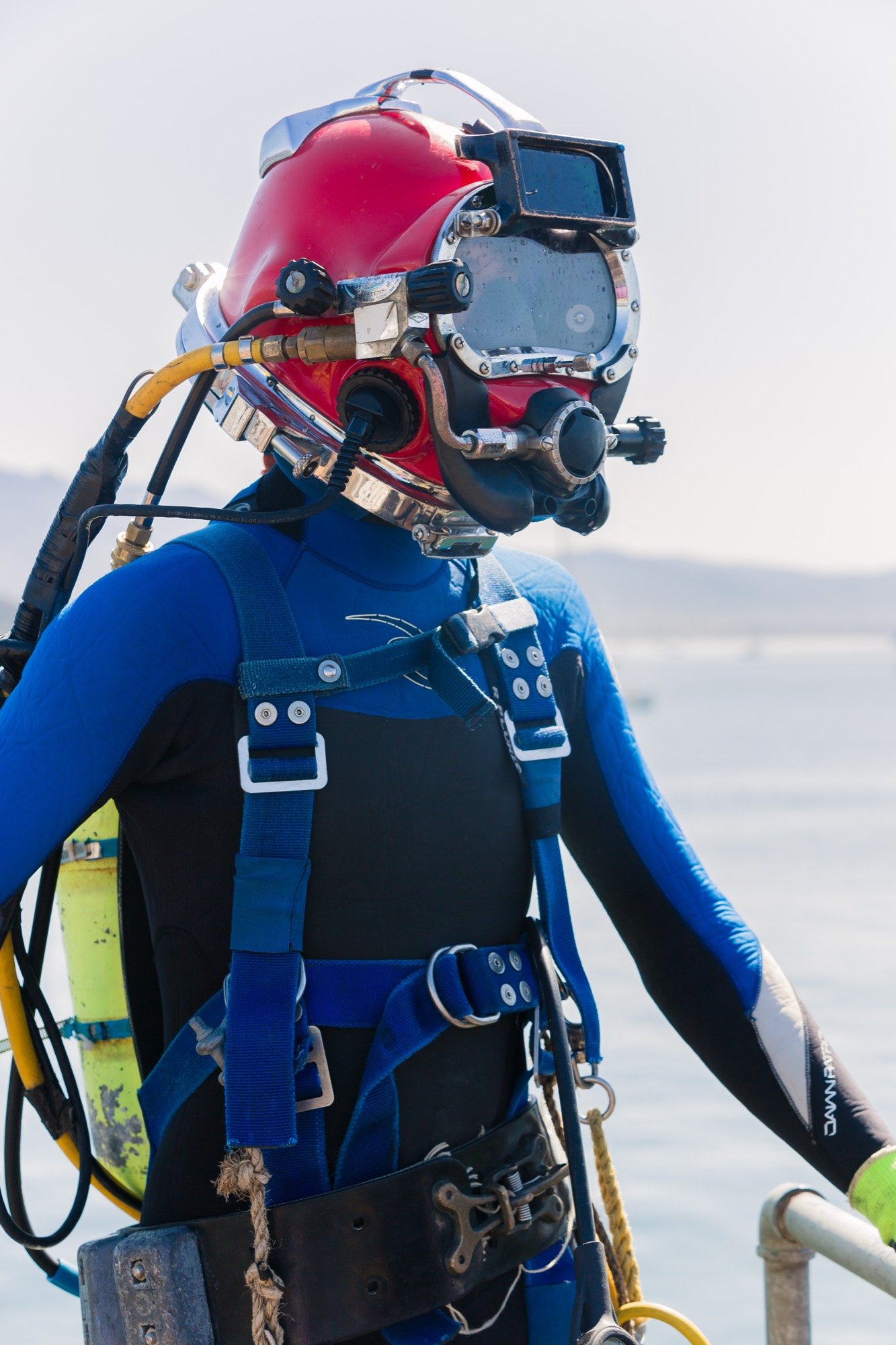Commercial diver in a red helmet and blue drysuit on a pier, ready to enter the water