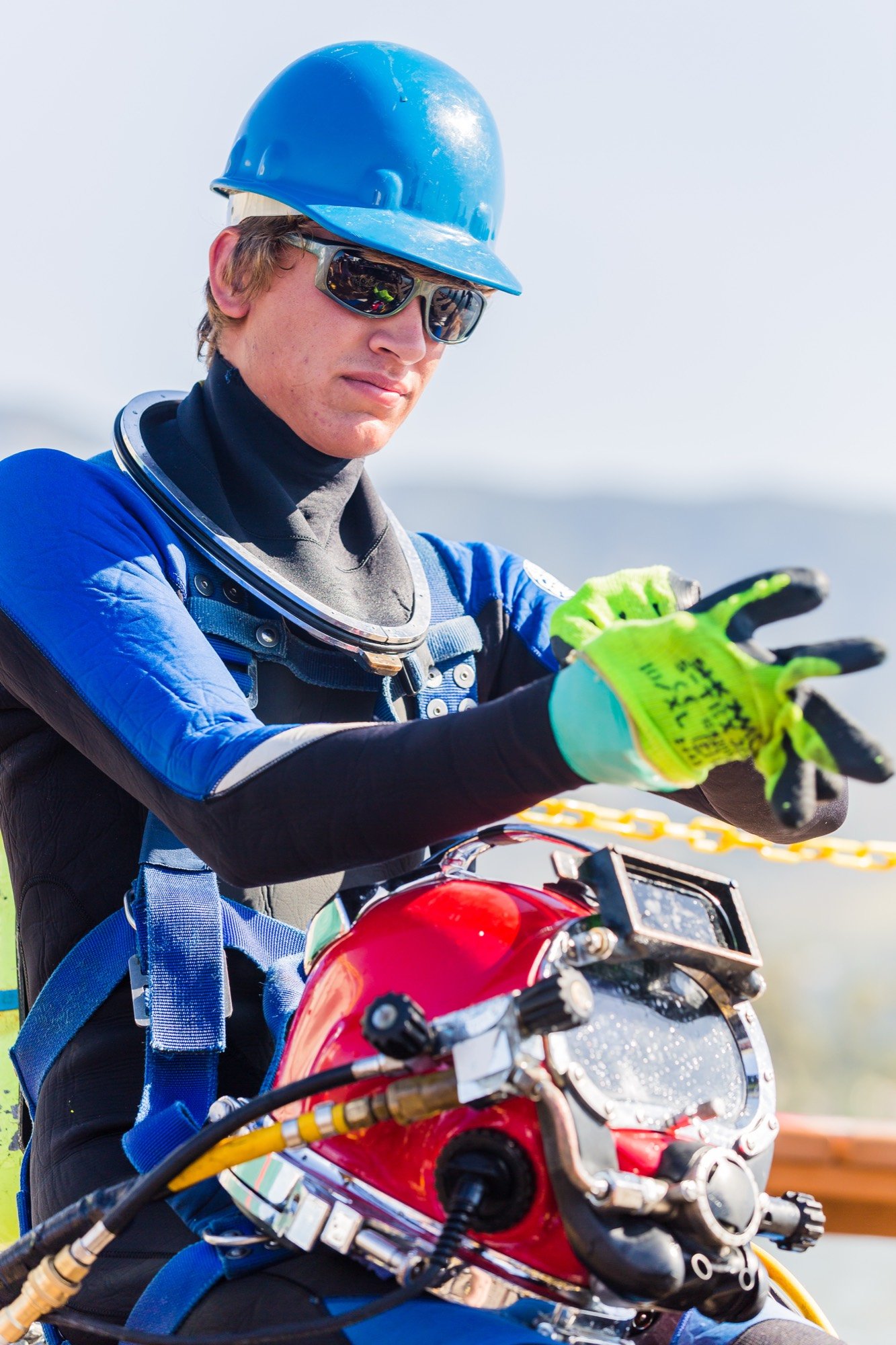 Diver in sunglasses and a hard hat checking a red dive helmet on his lap