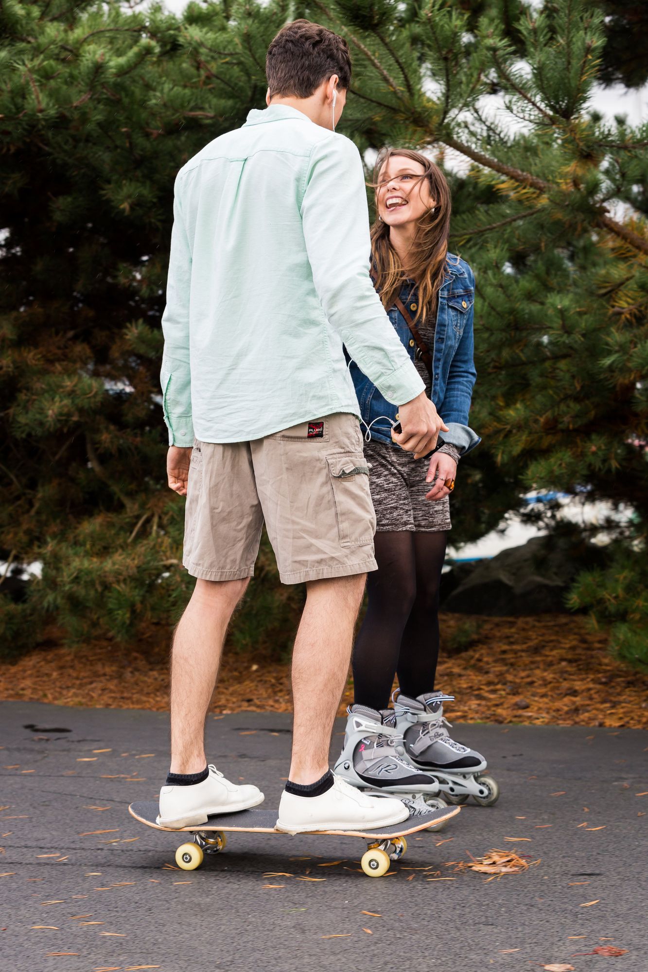 Young man on a skateboard and a laughing young woman on roller skates in a park under evergreen trees