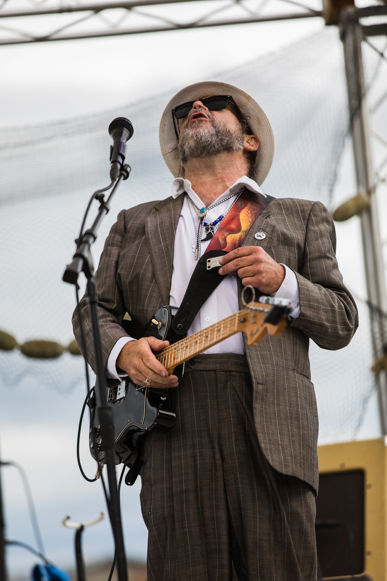 Guitarist in a fedora and pinstripe suit singing on an outdoor festival stage