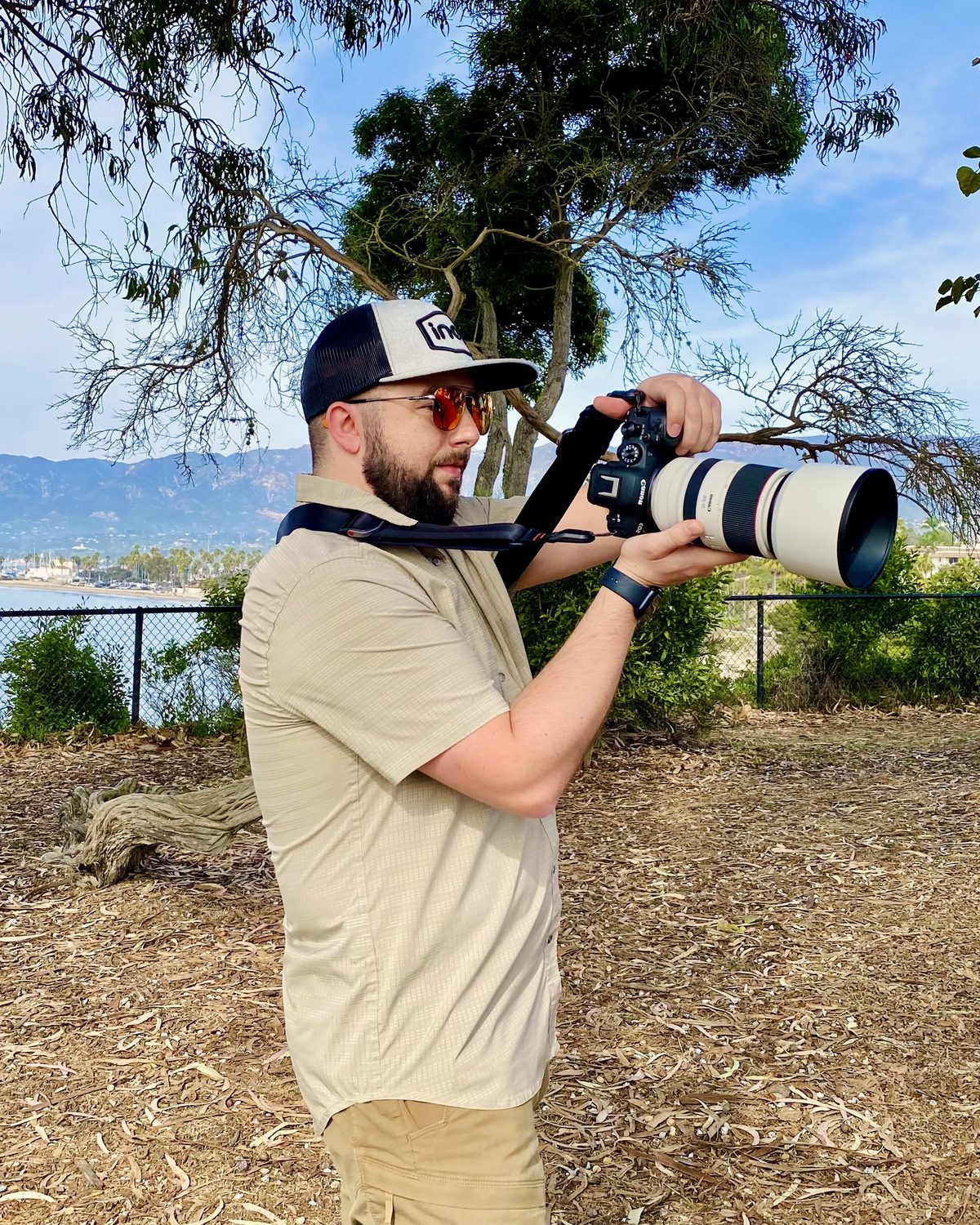 Gabriel Knapp in a cap and sunglasses photographing with a white telephoto lens on a bluff above Santa Barbara, the harbor and mountains behind him