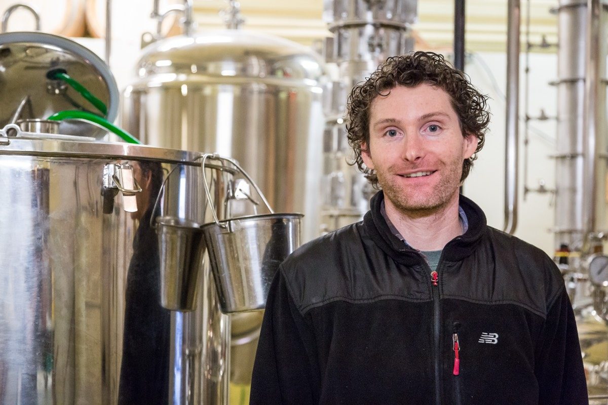 Smiling brewer in a black fleece among stainless steel brewing tanks