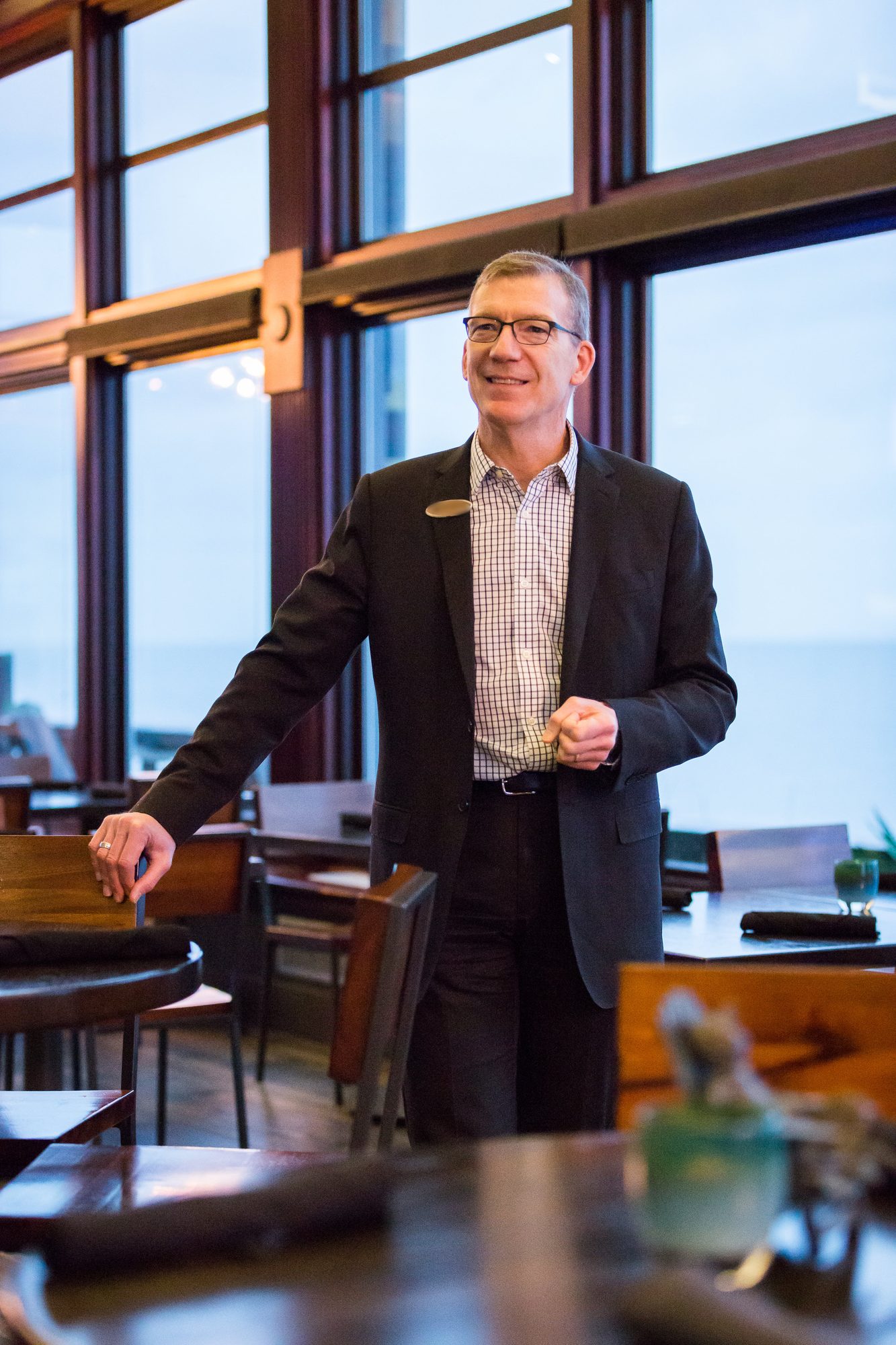 Executive in a dark suit leaning on a table in a sunlit restaurant with tall windows overlooking the water