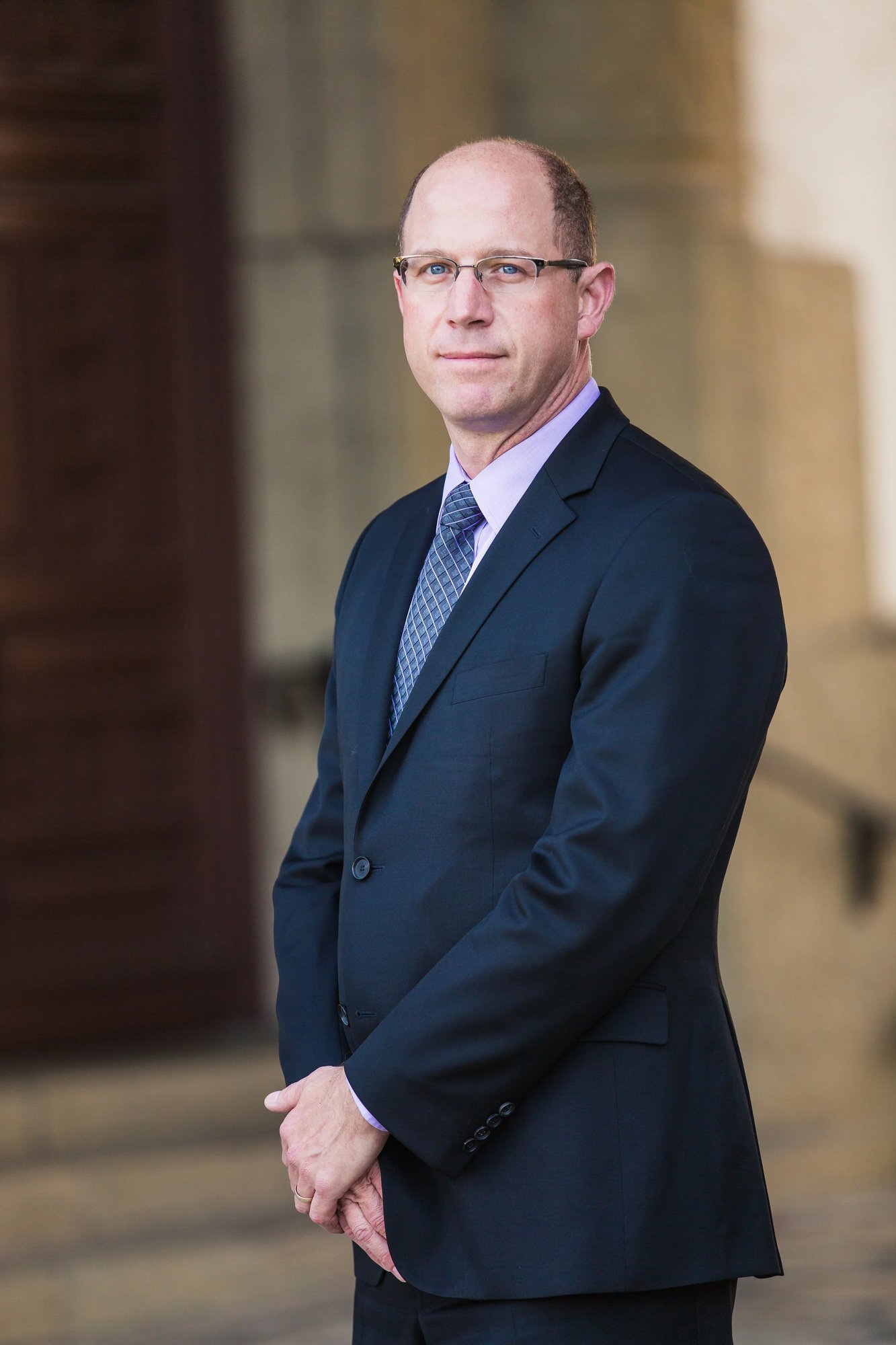 Corporate portrait of a bald executive in glasses and a navy suit standing beside a stone column