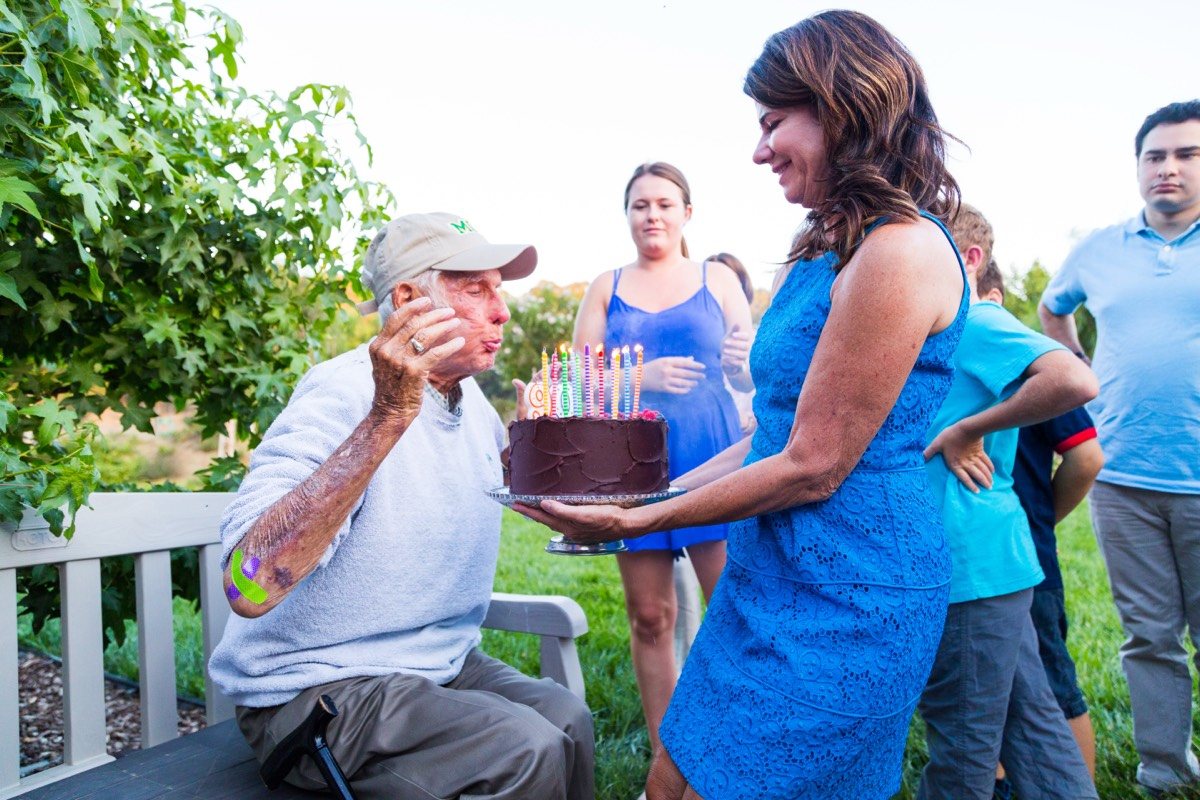 Older man in a sun hat blowing out the candles on a chocolate birthday cake held by a woman in a blue dress, with guests gathered in a garden