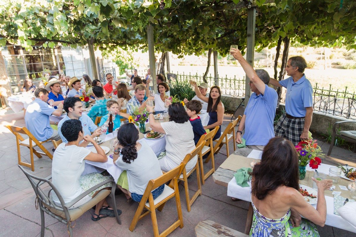 Long dinner table of party guests under a grapevine pergola, one guest standing with a raised arm