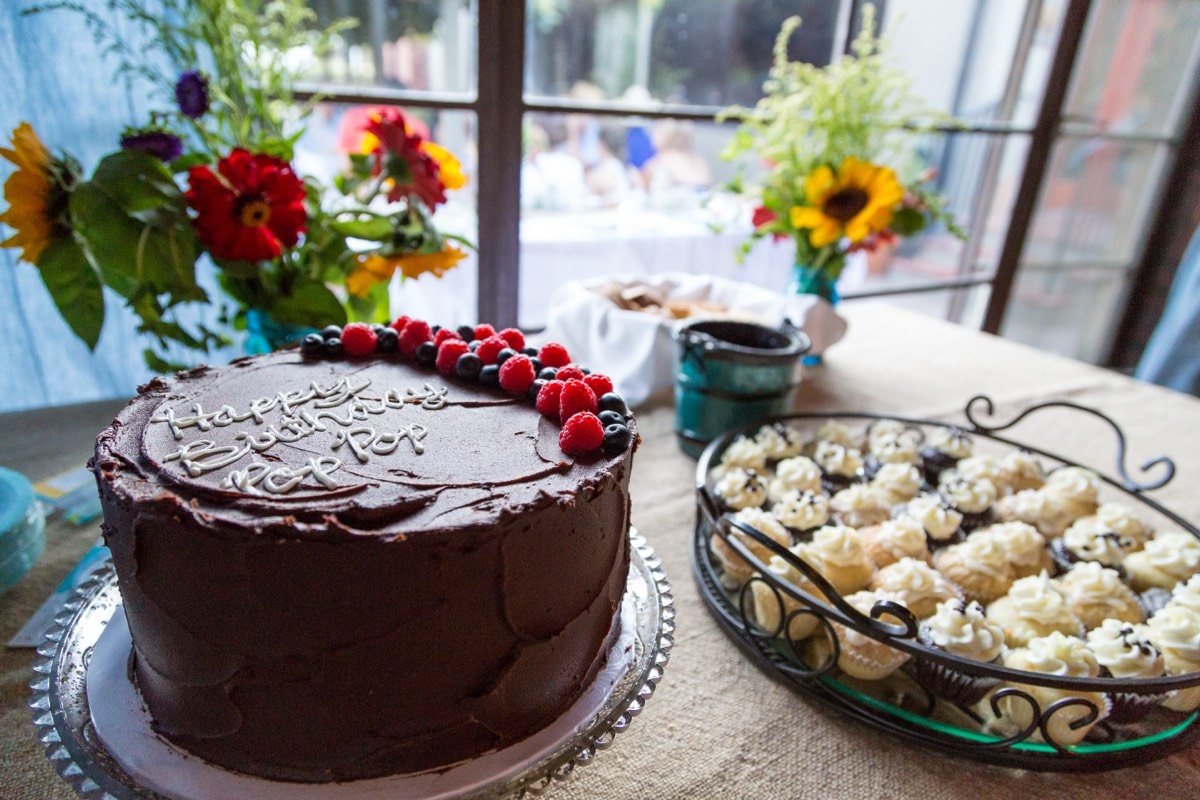 Chocolate birthday cake topped with berries beside flowers and a tray of pastries