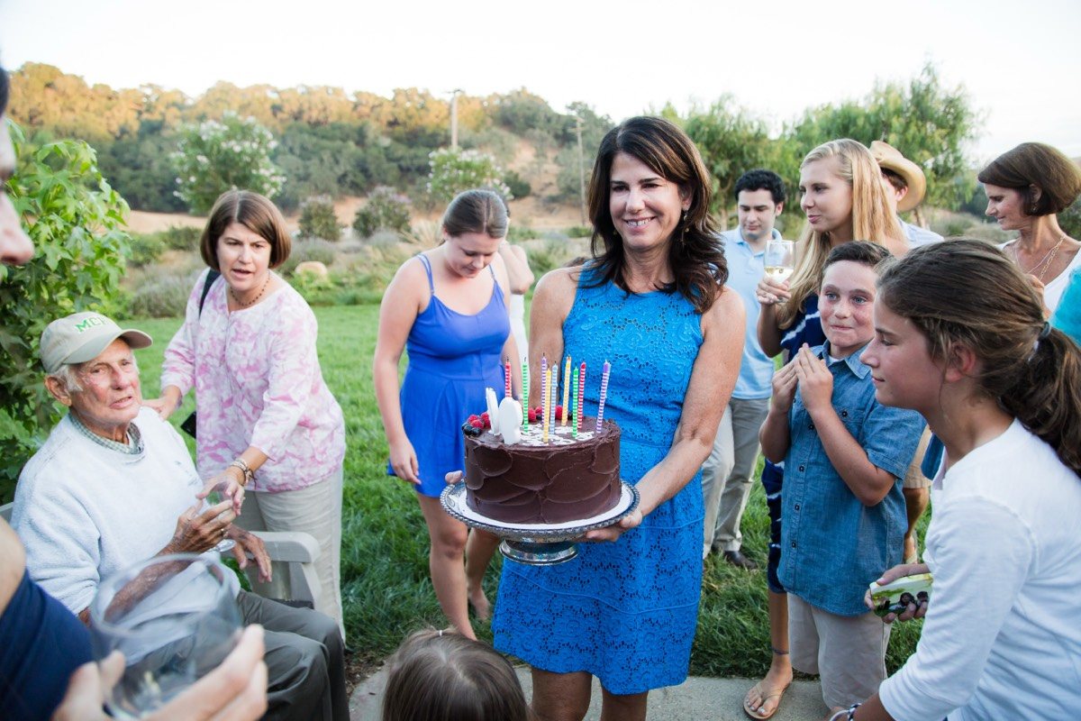Woman in a blue dress carrying a candlelit birthday cake through smiling guests on a lawn