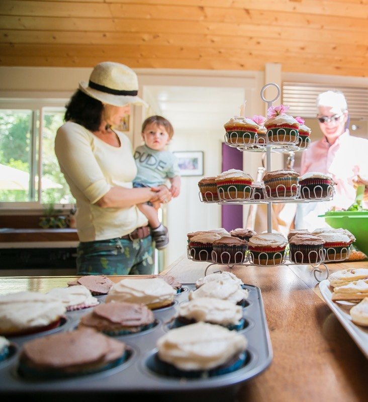 Woman holding a toddler beside a cupcake stand and trays of pastries in a kitchen