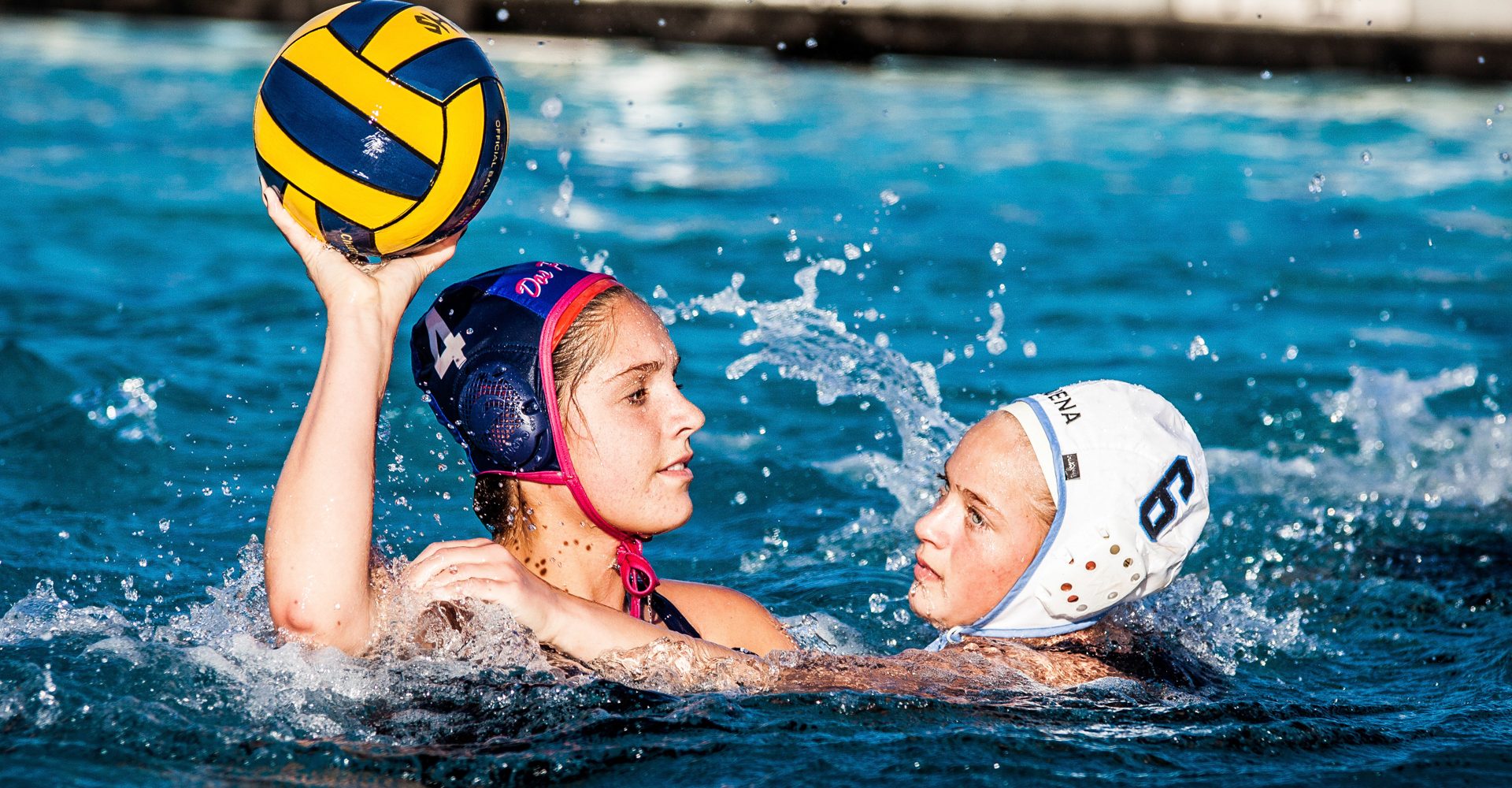 Water polo player raising the ball to shoot as a defender closes in, spray in the air