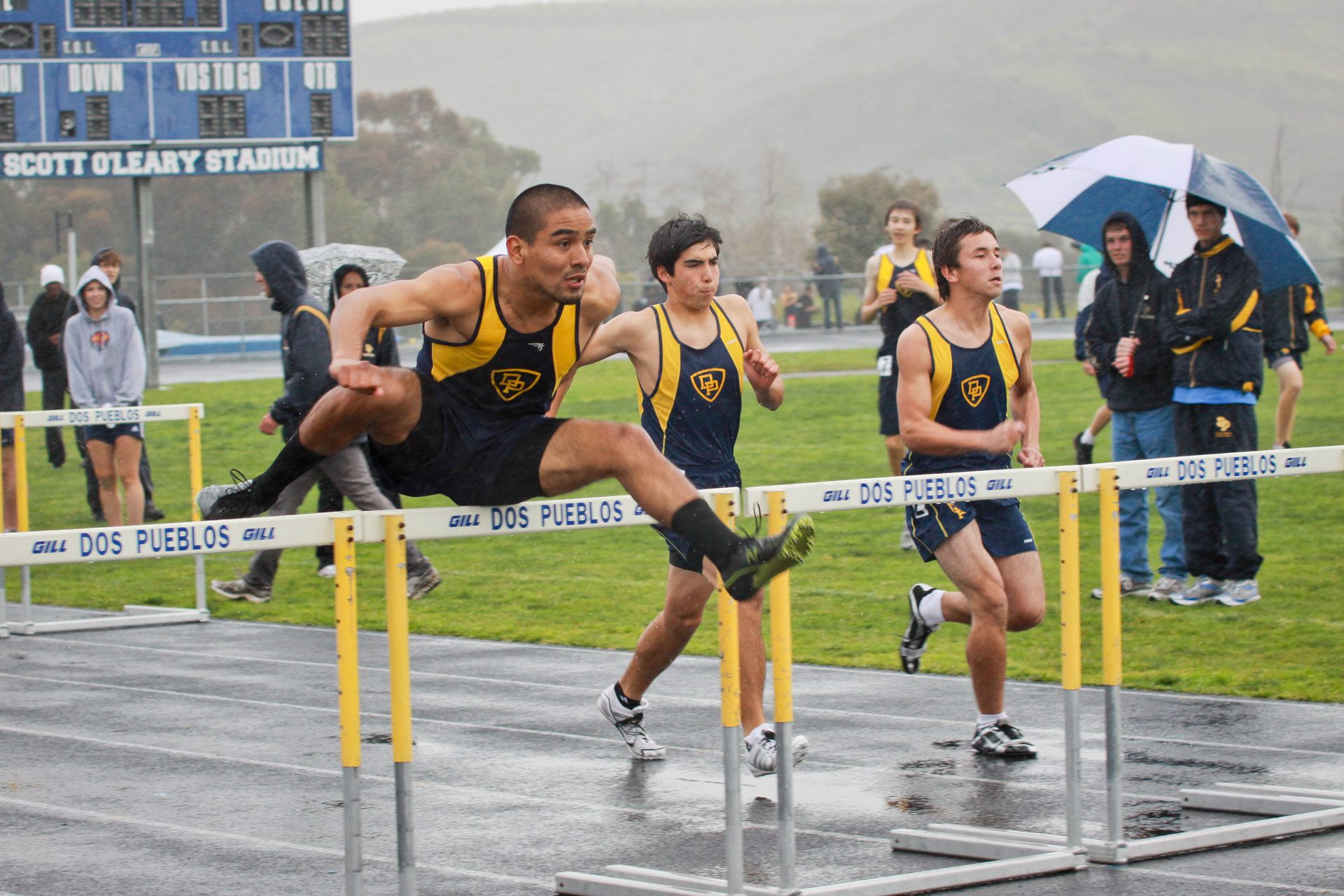 Two hurdlers clearing a barrier side by side on a wet track meet